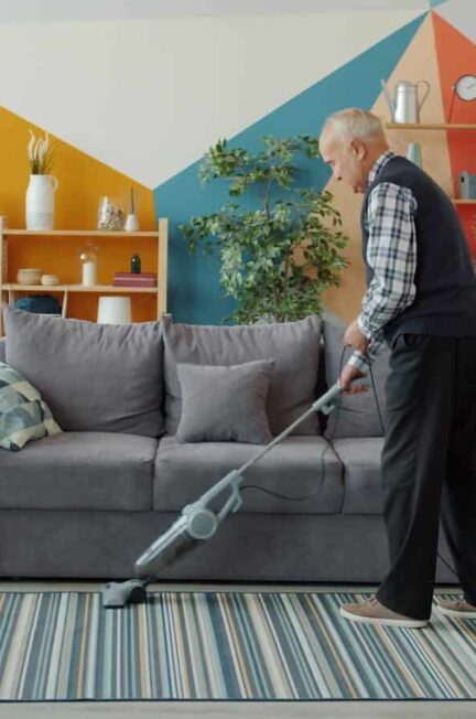 Elderly woman vacuuming a colorful living room floor.