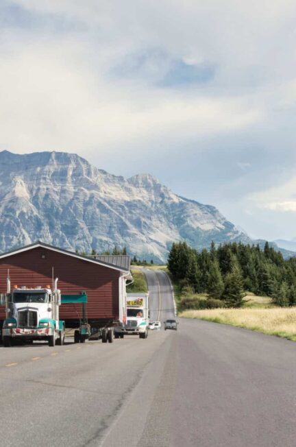Semi-trucks transporting mobile homes on a rural road with mountain backdrop.