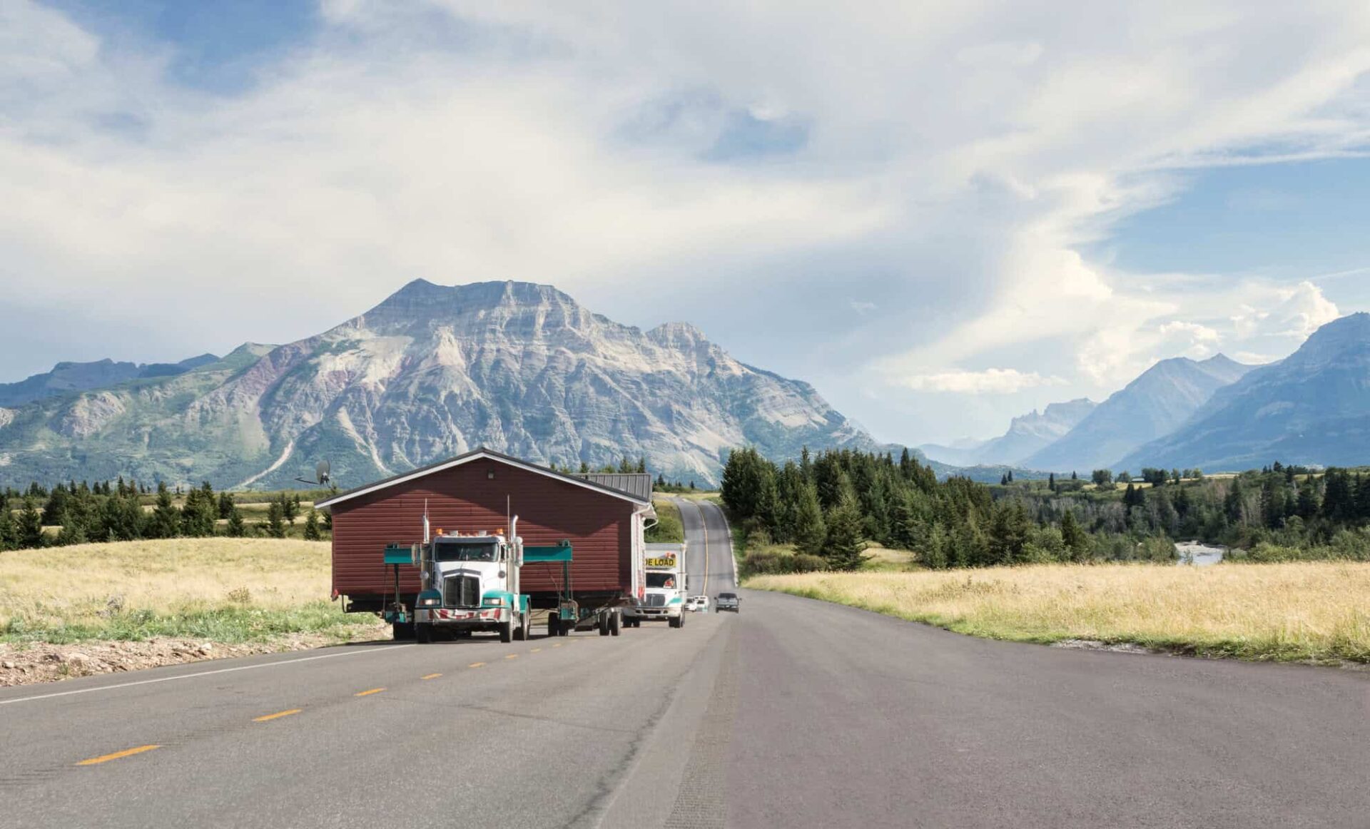 Semi-trucks transporting mobile homes on a rural road with mountain backdrop.