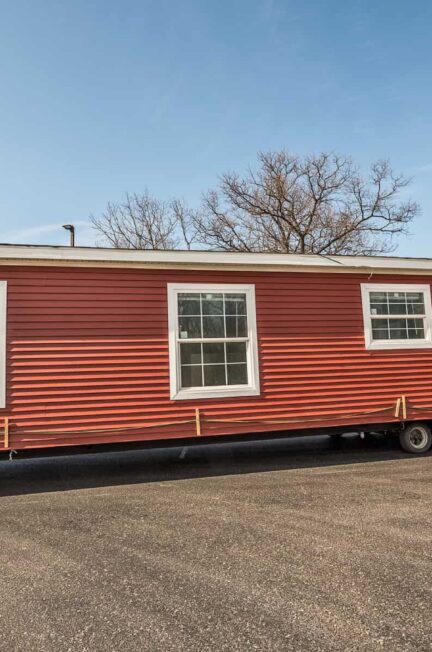Red mobile home on a paved lot with trees in the background.