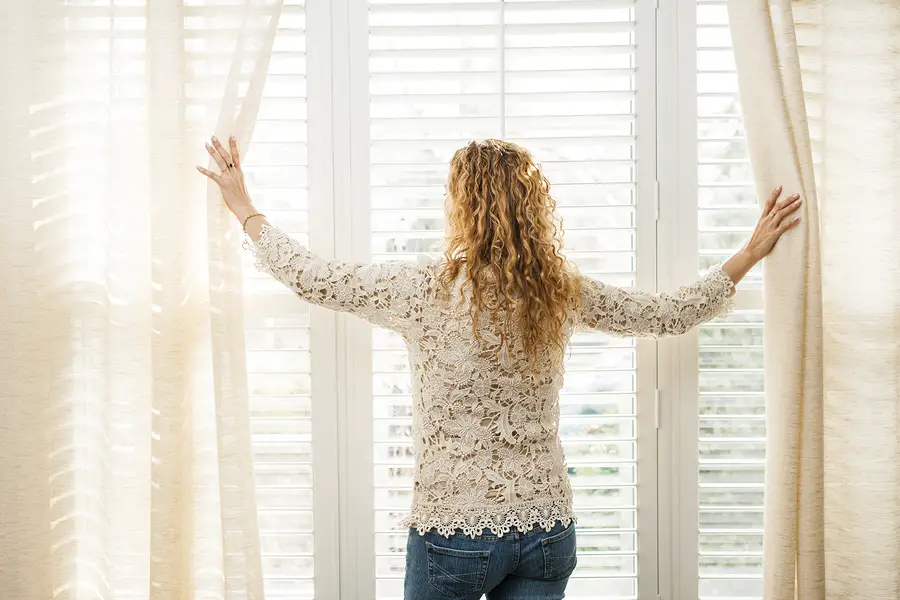 Woman looking out big bright window with curtains and blinds